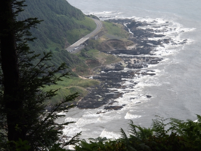 [Irregular-surfaced dark rock sits between the white-capped waves of the ocean and the greenery of the hillside just below the roadway.]